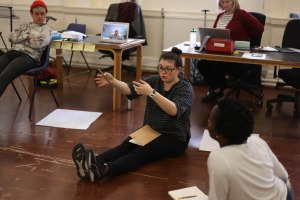 Eurasian woman with dark hair in a high bun and wearing glasses is sat on the wooden floor of a rehearsal room with her legs outstretched. She has a notebook in her lap and is gesticulating with both hands. There are tables in the background and a black woman is sat listening in the foregrount.