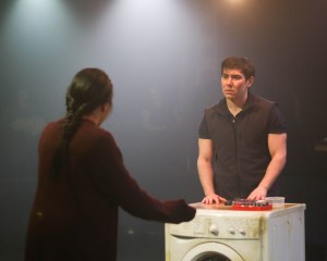 A young Eurasian man stood behind a grubby-looking washing machine talking to an East Asian woman who has her back to us.