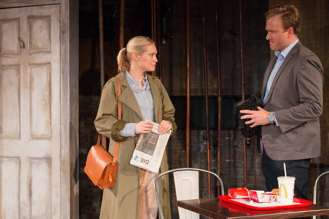 A blonde woman wearing a long khaki raincoat and carrying a brown leather shoulder bag, holding a German newspaper in her hands talks to a blonde man in a tweed jacket. There is a red tray on the table with the remnants of a fast food meal.
