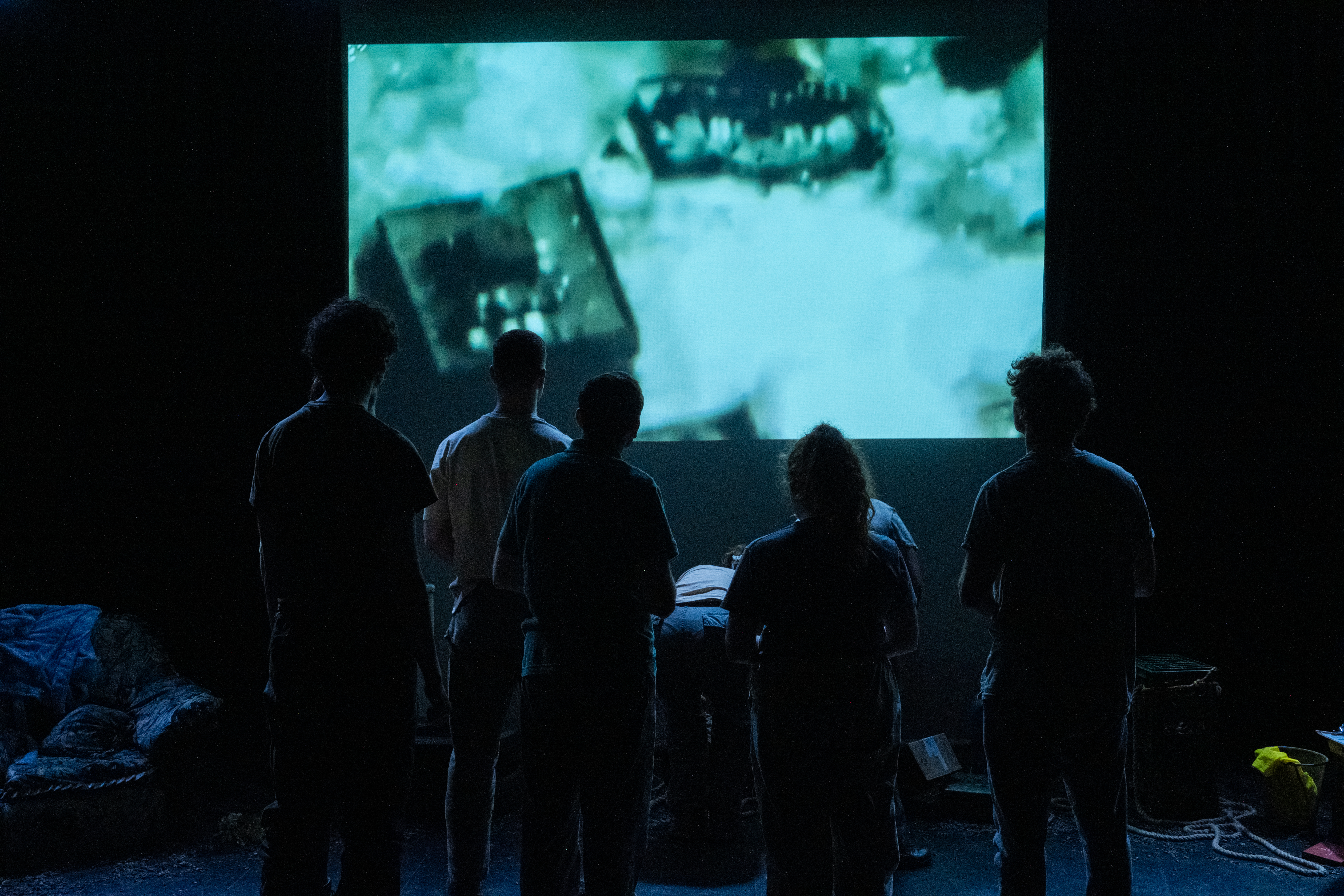 Image from a theatre production where actors playing the citizens are watching a film of a bomb exploding. We see them in silhouette against the projected image.