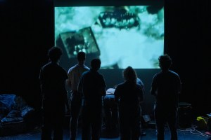 Image from a theatre production where actors playing the citizens are watching a film of a bomb exploding. We see them in silhouette against the projected image.